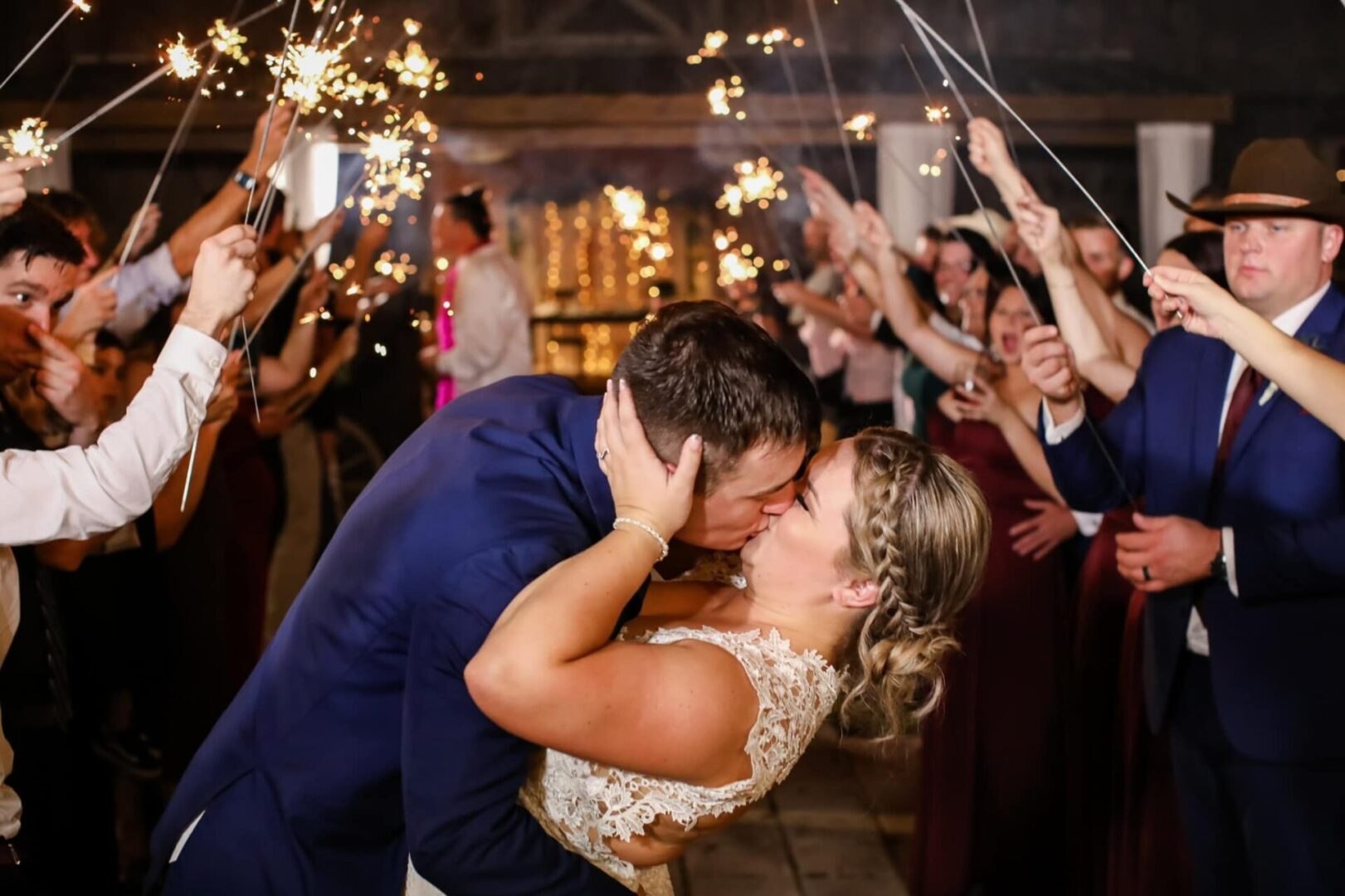 Newlyweds share a passionate kiss surrounded by cheering guests with sparklers.