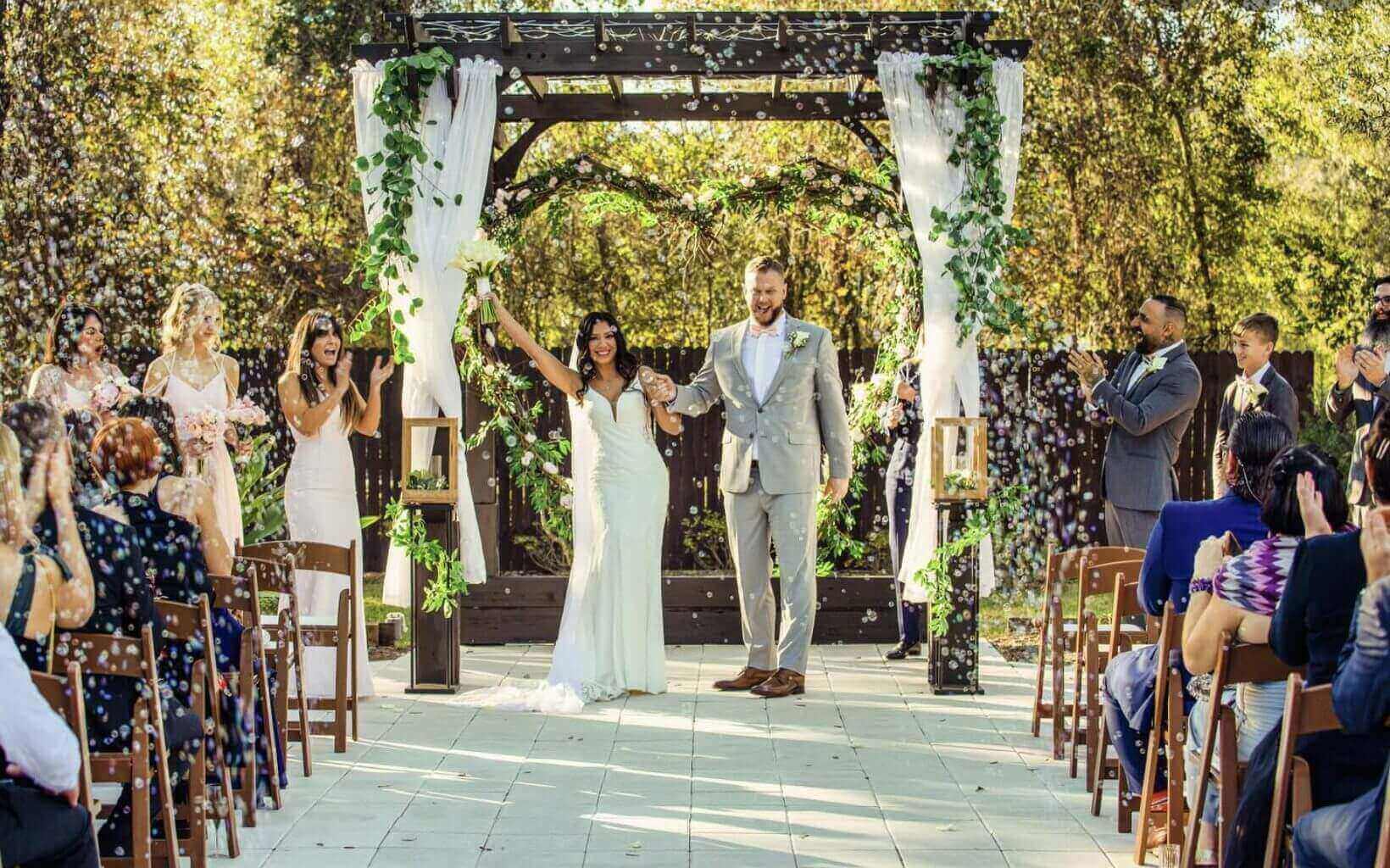Newlyweds joyfully exit their outdoor wedding ceremony under a floral arch.