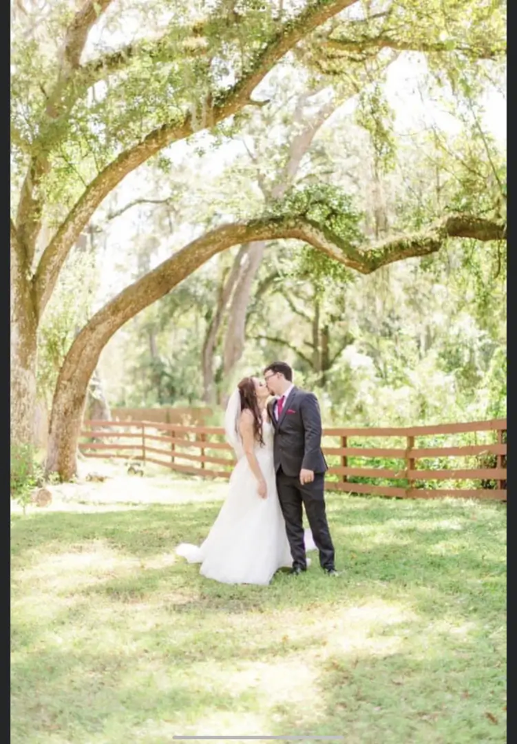 Bride and groom share a kiss under a large tree in a sunlit garden.